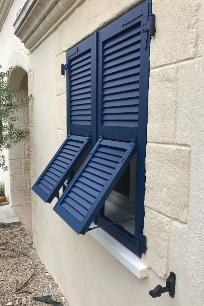 Volets battants persiennés bleu marine partiellement ouverts sur une façade en pierre claire, avec une petite fenêtre et un arrêtoir noir.