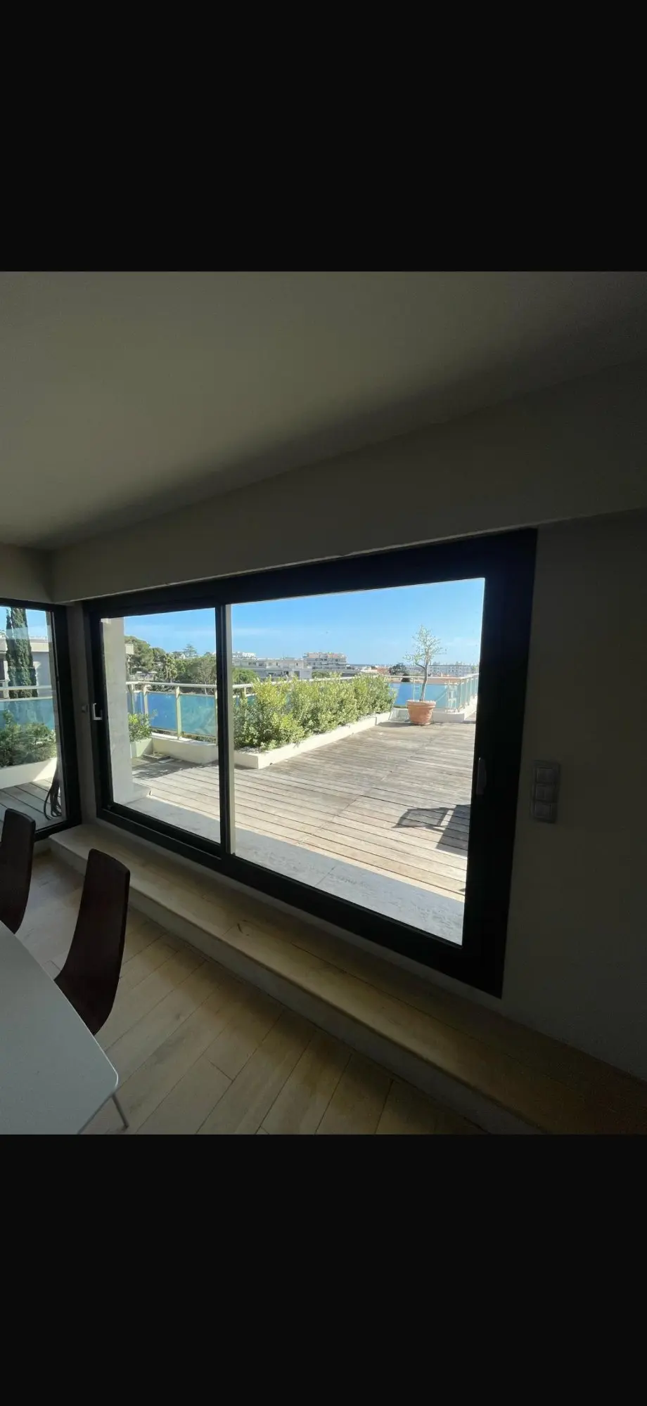 Intérieur moderne avec sol en bois clair. Vue à travers la grande baie vitrée sur une vaste terrasse en bois, végétation et bâtiments côtiers sous ciel bleu.