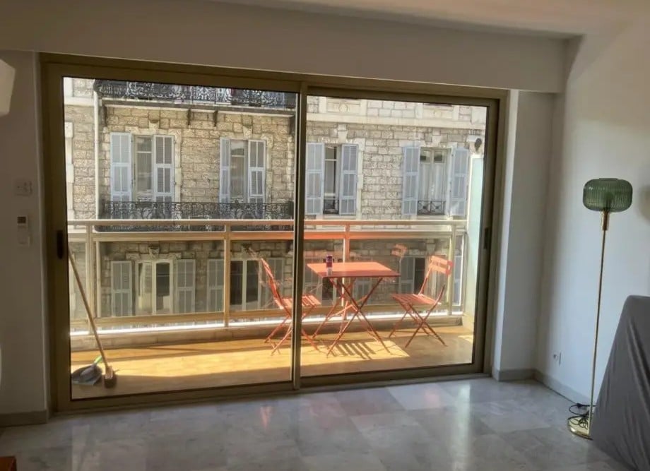 Intérieur lumineux, sol en marbre. Balcon en bois avec table bistro orange. Vue urbaine sur façade historique en pierre brute et volets gris.