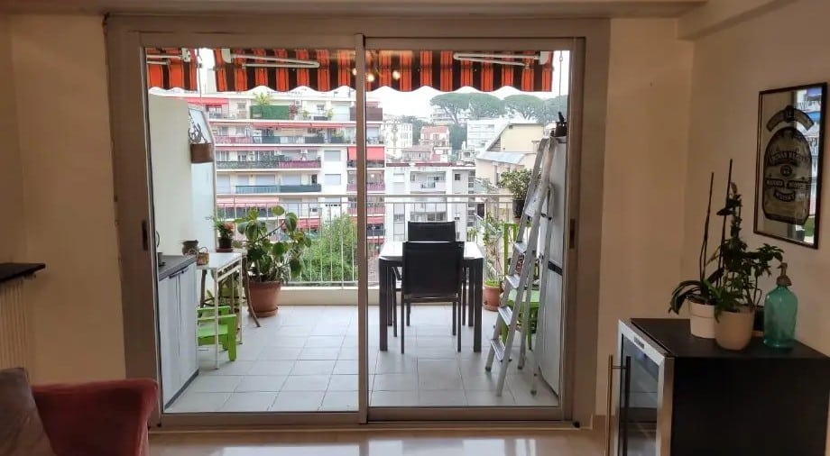 Intérieur lumineux donnant sur un grand balcon avec table à manger, plantes et auvent rayé, offrant une vue panoramique sur une ville méditerranéenne.
