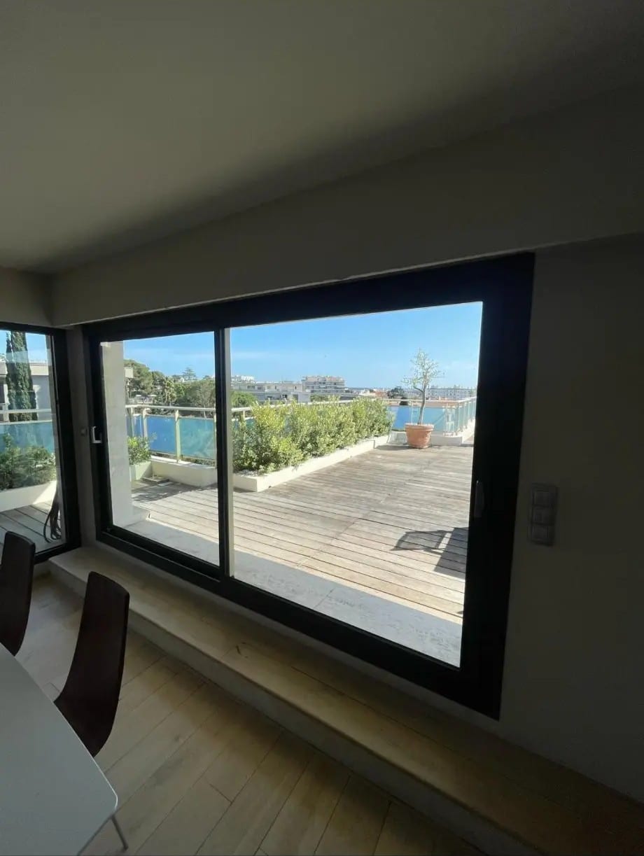 Intérieur moderne avec vue imprenable à travers une baie vitrée noire sur une grande terrasse en bois, ciel bleu et jardin méditerranéen.