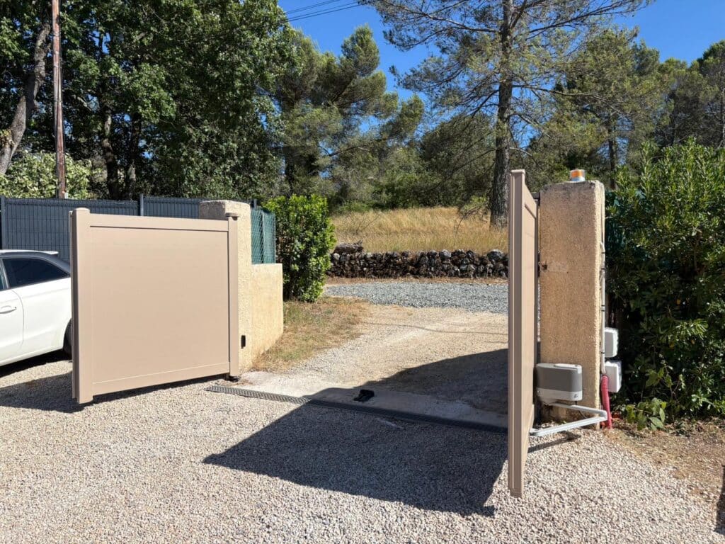Portail battant automatique en aluminium beige ouvert sur une allée en gravier. Moteur fixé au pilier. Clôture verte et arbres en arrière-plan.