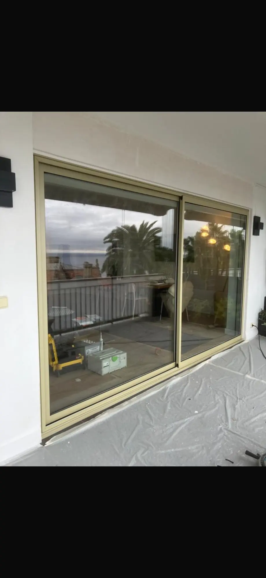 Porte coulissante vitrée moderne, cadre alu beige. Balcon en rénovation, sol protégé. Reflet d'outils, palmier et vue mer sous ciel nuageux.
