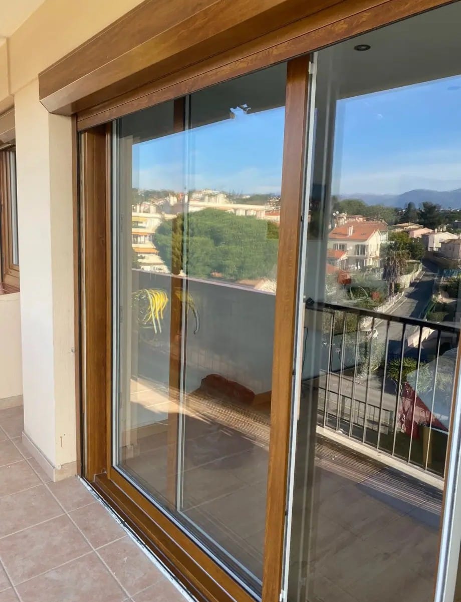 Grande porte coulissante vitrée aspect bois menant à un balcon. Système de volet roulant supérieur visible. Vue ensoleillée sur la ville.