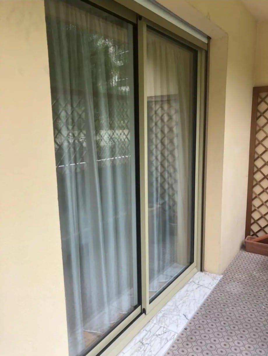 Grande porte coulissante en aluminium beige. Un voilage blanc et une grille de sécurité sont visibles. Terrasse avec carrelage géométrique et marbre.