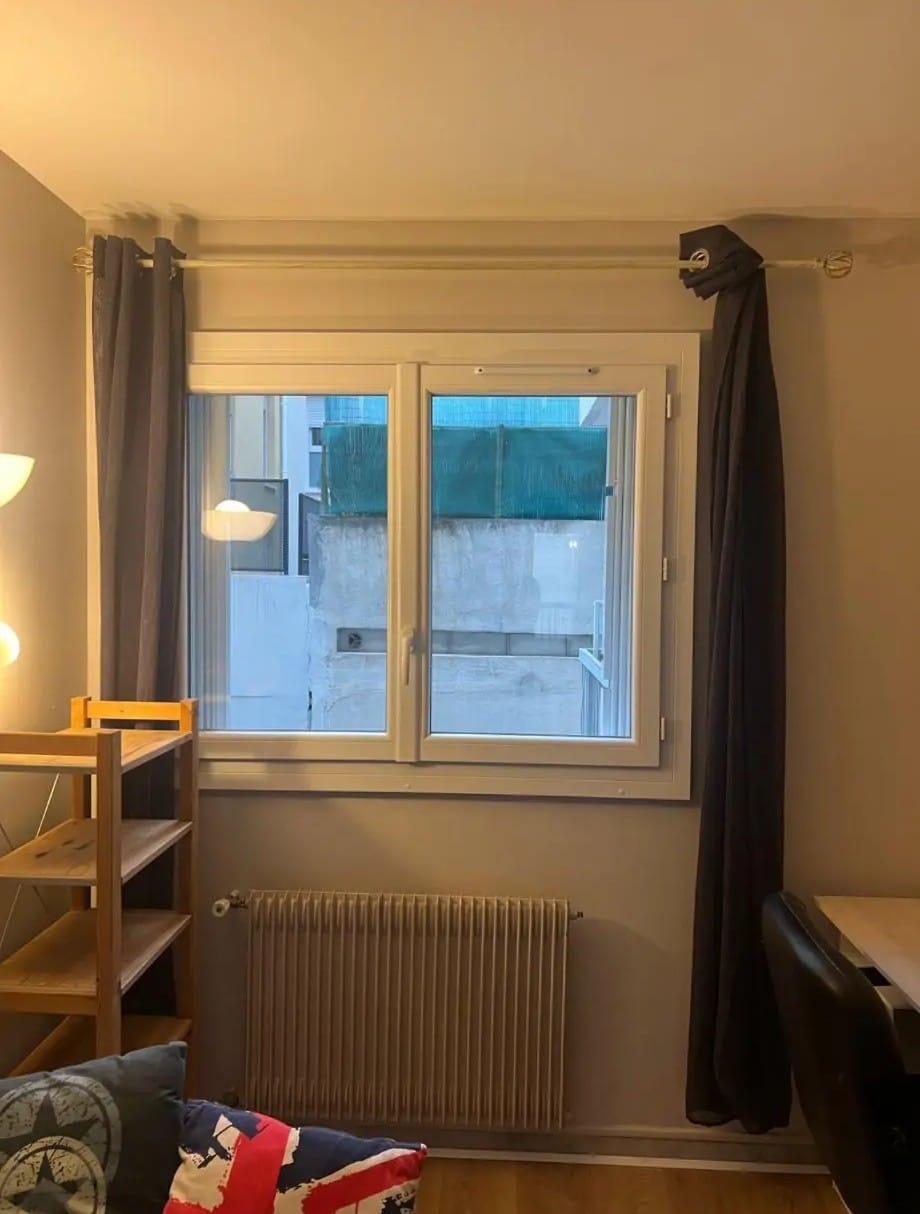 Salon urbain: fenêtre double avec vue sur cour, radiateur, rideaux gris occultants et étagère en bois. Coussins Union Jack en premier plan.