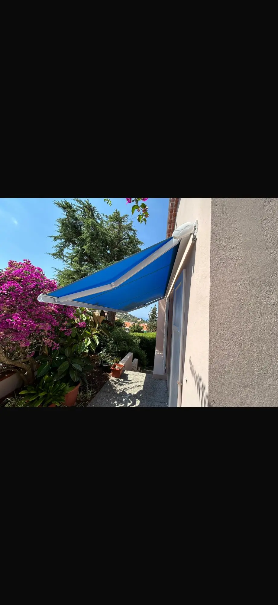 Store banne extérieur rétractable bleu sur mur beige, protégeant une terrasse. Jardin fleuri, bougainvillier pourpre et vue mer au loin.