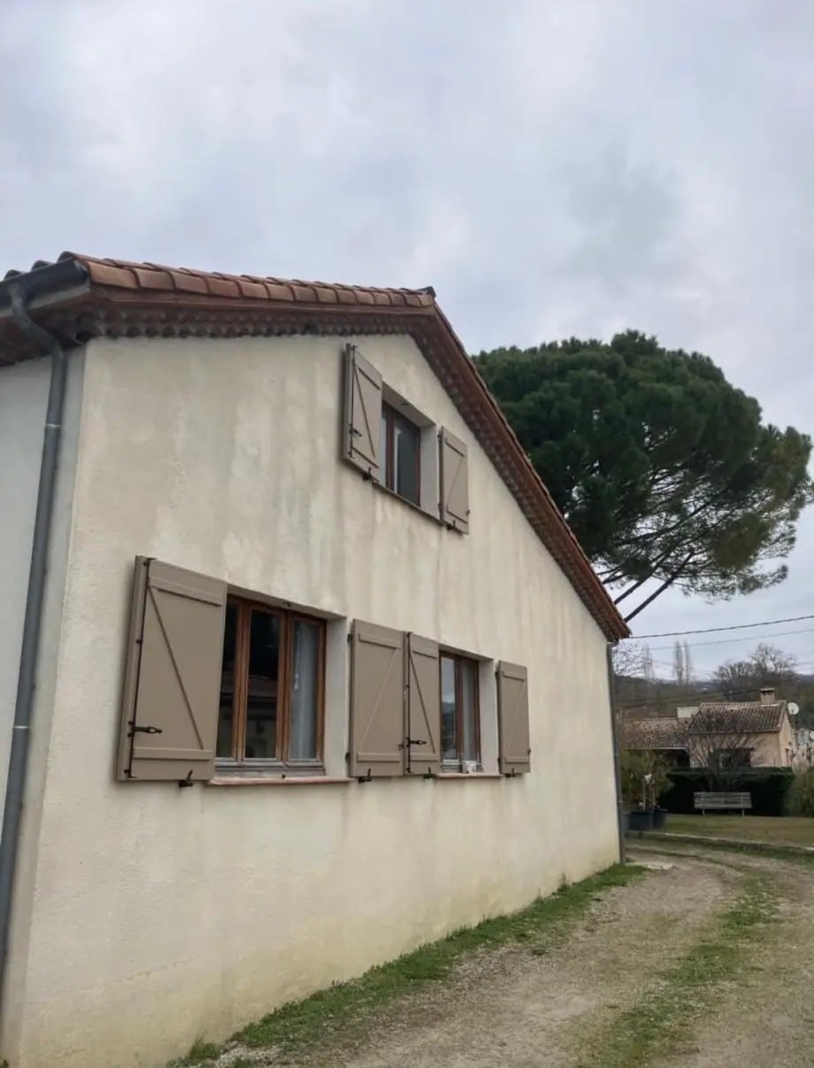 Façade beige d'une maison rurale provençale avec des volets battants taupe et un toit en tuiles de terre cuite sous un ciel gris.