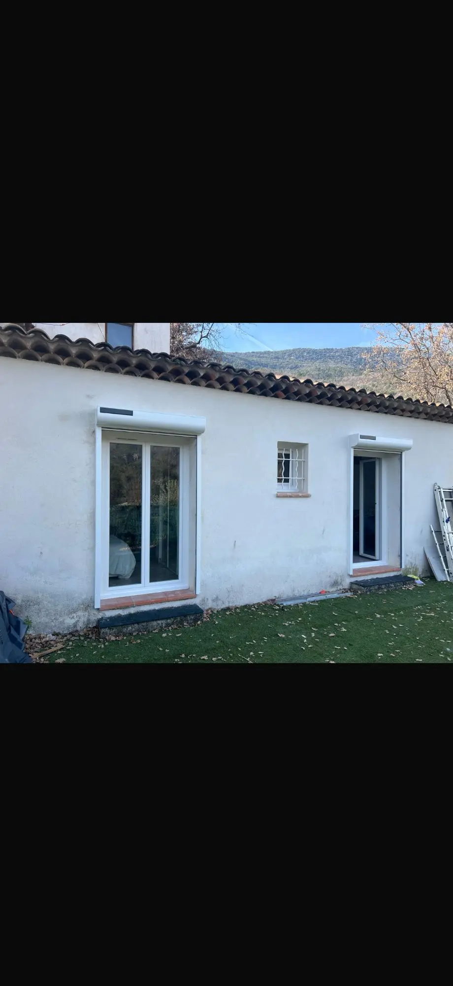 Façade maison blanche rénovée, deux portes-fenêtres PVC équipées de volets roulants. Toit tuiles traditionnelles, petit jardin gazonné.