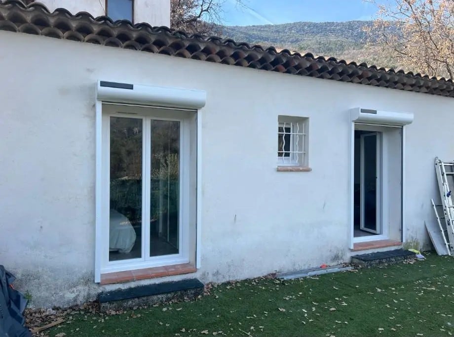 Façade blanche en rénovation avec deux portes-fenêtres et un judas à barreaux. Volets roulants blancs apparents, toit en tuiles romanes.