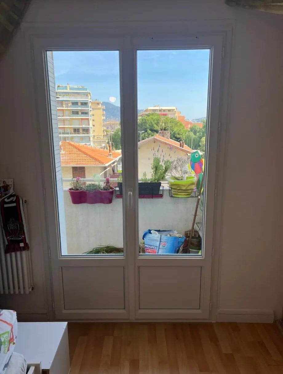 Intérieur lumineux avec vue sur la ville depuis une porte-fenêtre blanche. On voit des toits, des immeubles et un balcon fleuri. Écharpe OGC Nice.