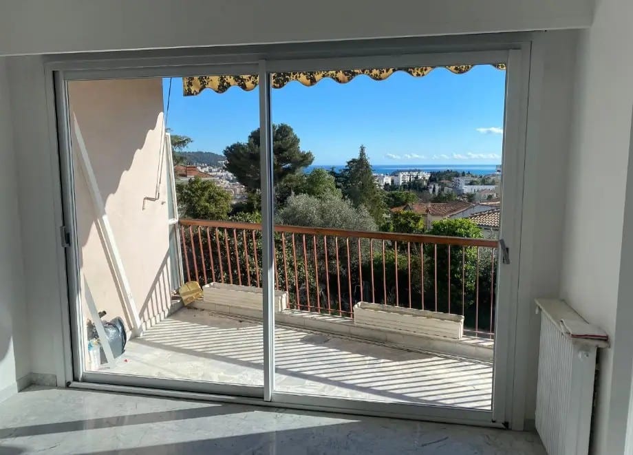 Baies vitrées coulissantes ouvrant sur un balcon avec rambarde. Vue mer panoramique sur la côte et les toits, entourée de verdure sous un ciel bleu éclatant.