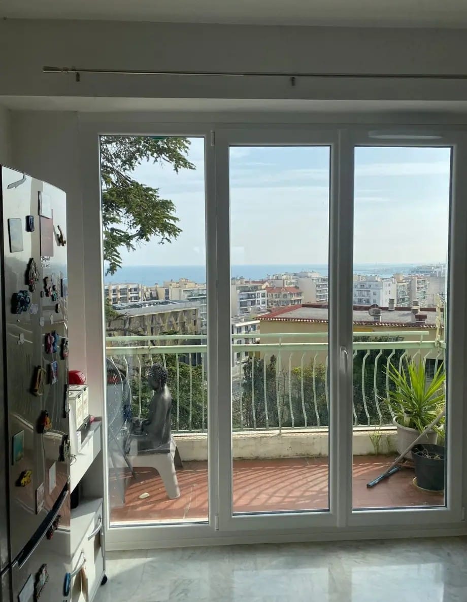 Vue mer et ville côtière depuis un intérieur. Grande porte-fenêtre PVC donne sur un balcon ensoleillé avec statue et garde-corps.